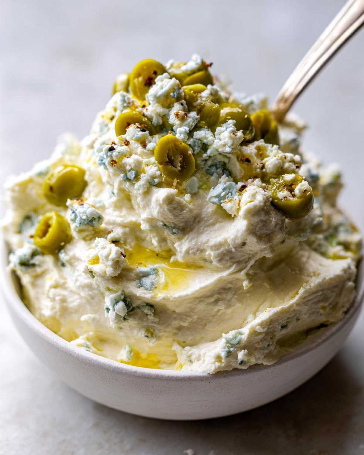 A close-up of creamy dirty martini dip topped with sliced green olives, blue cheese crumbles, and olive oil.