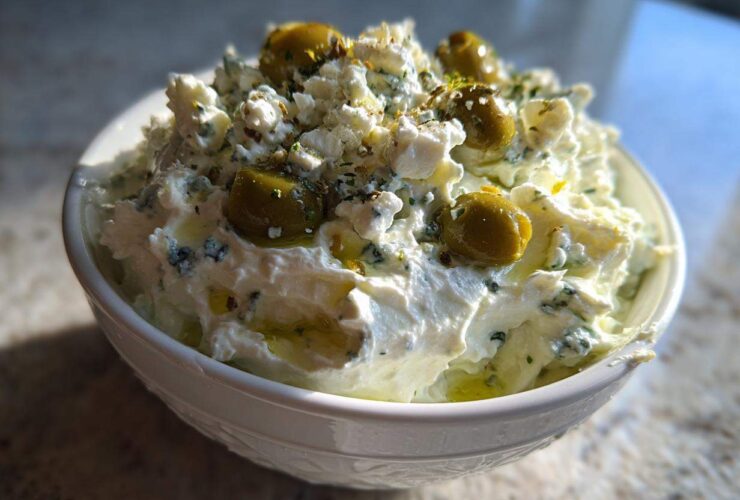 Close-up of a creamy, white dirty martini dip topped with crumbled cheese, green olives, and herbs.