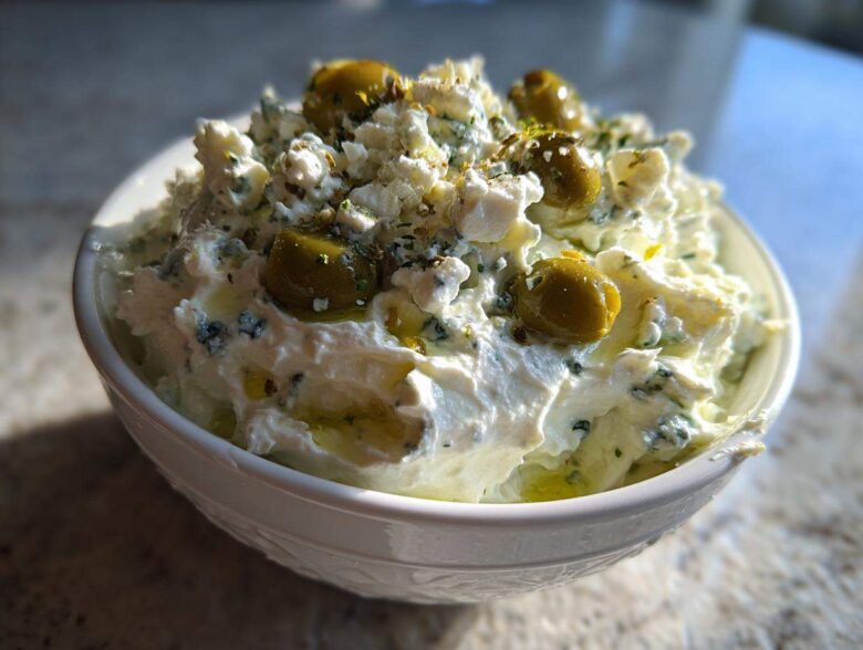Close-up of a creamy, white dirty martini dip topped with crumbled cheese, green olives, and herbs.