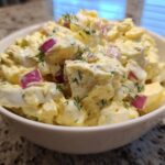 Close-up of a white bowl filled with creamy egg salad recipe, featuring chunks of egg, red onion, and dill.