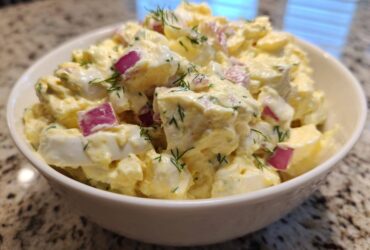 Close-up of a white bowl filled with creamy egg salad recipe, featuring chunks of egg, red onion, and dill.