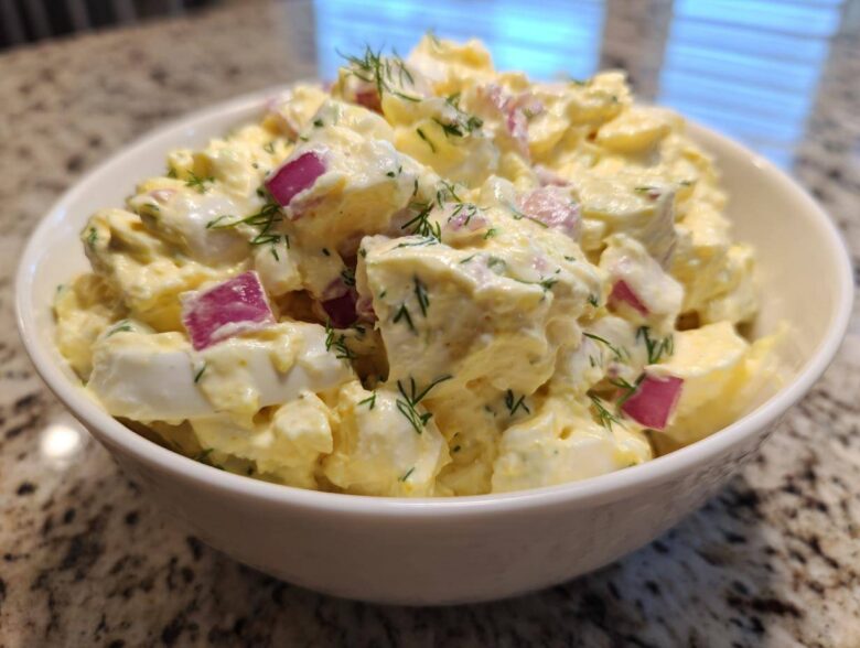 Close-up of a white bowl filled with creamy egg salad recipe, featuring chunks of egg, red onion, and dill.