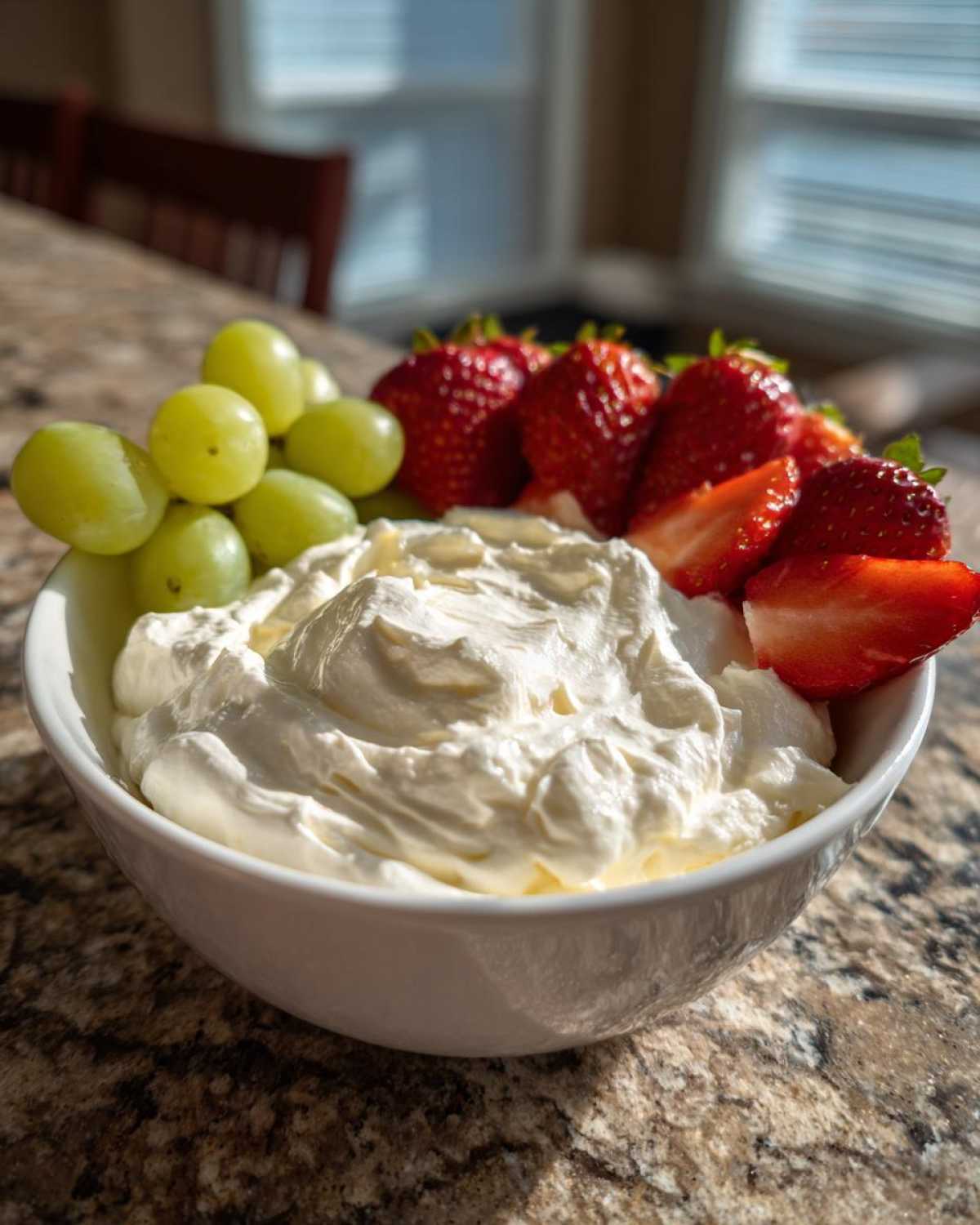 A white bowl filled with creamy fruit dip, surrounded by fresh green grapes and bright red strawberries.