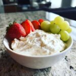 A white bowl filled with creamy fruit dip, garnished with fresh strawberries and green grapes.