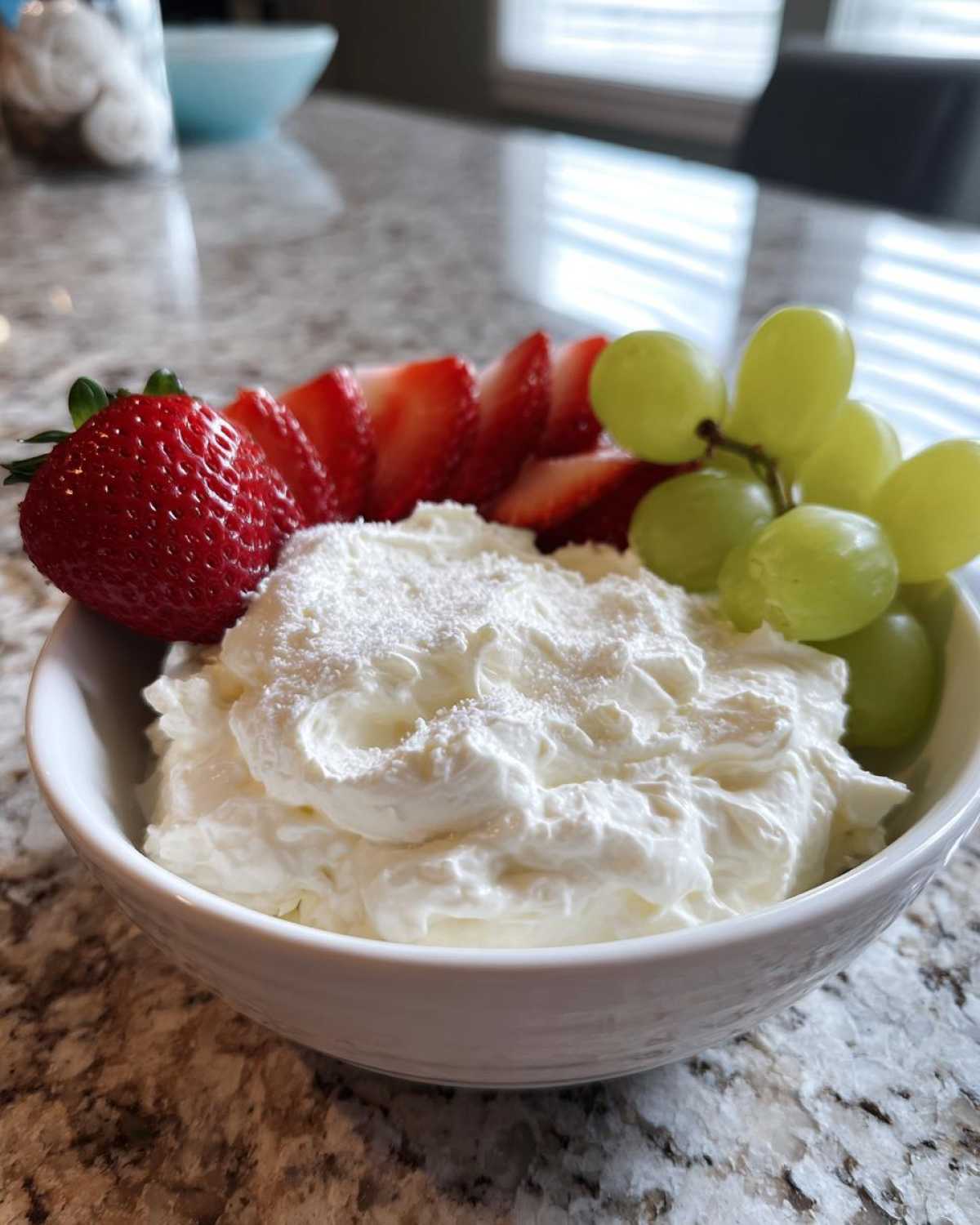 White bowl filled with creamy fruit dip, garnished with sliced strawberries and green grapes.