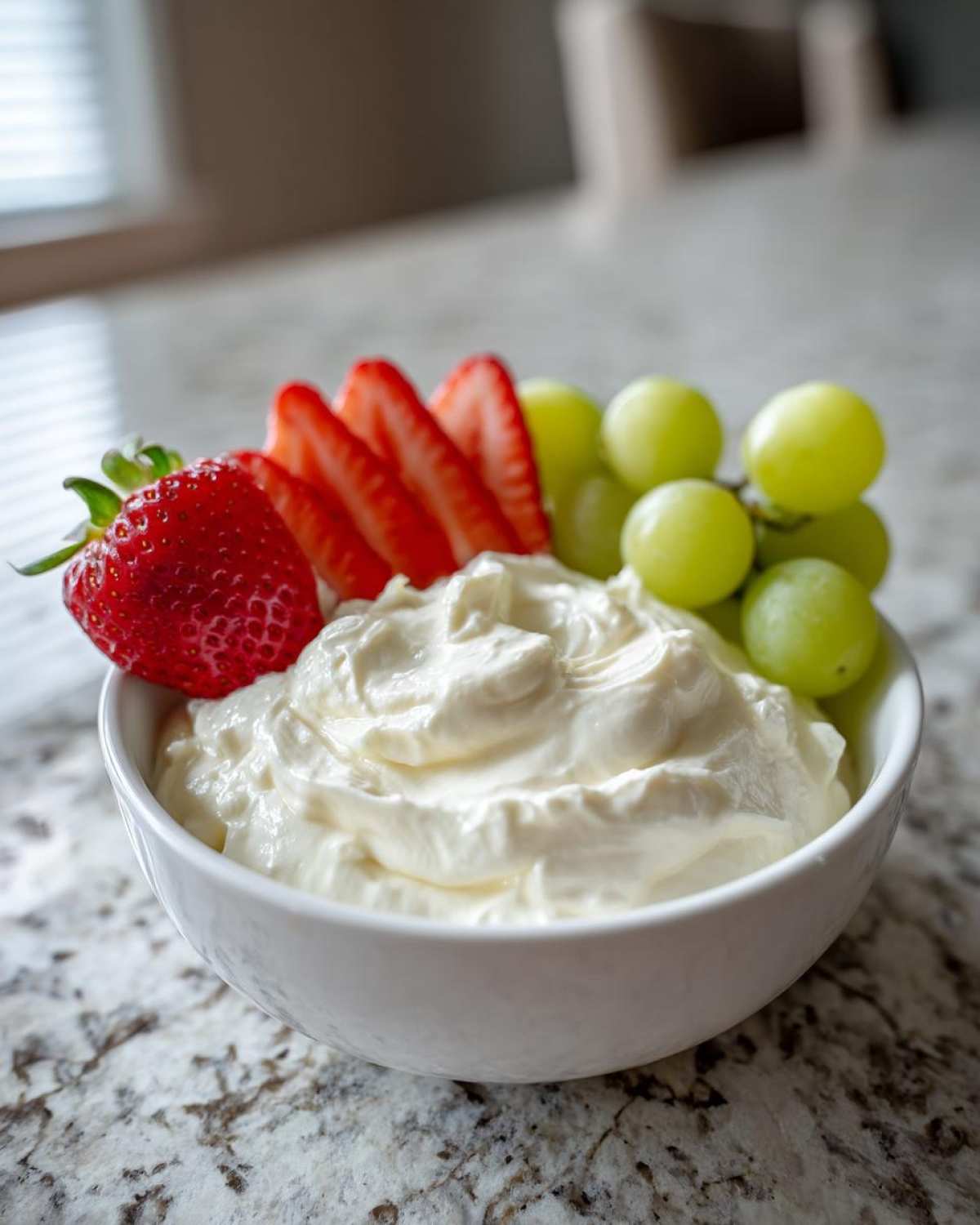 A white bowl filled with creamy fruit dip, garnished with sliced strawberries and green grapes.