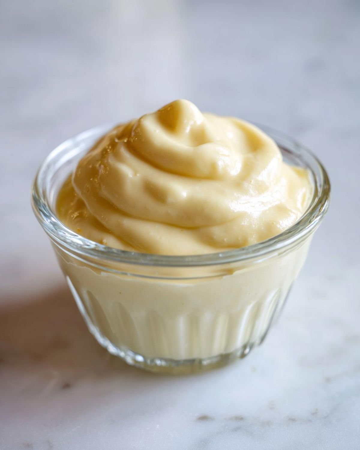 A close-up of thick, creamy homemade mayonnaise swirled high in a small glass ramekin on a white marble surface.