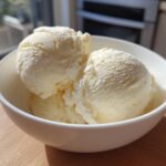 Three scoops of creamy, light yellow protein ice cream served in a white bowl on a wooden surface.