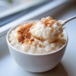 A close-up of creamy rice pudding topped with cinnamon, with a spoonful being lifted from the white bowl.
