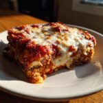A close-up, sunlit slice of crispy eggplant parmesan layered with rich tomato sauce and melted mozzarella cheese.