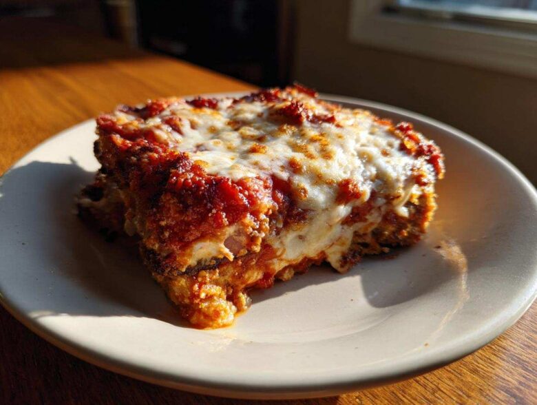 A close-up, sunlit slice of crispy eggplant parmesan layered with rich tomato sauce and melted mozzarella cheese.