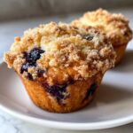 A close-up of a golden brown blueberry muffin topped with sweet, crunchy streusel, sitting on a white plate.