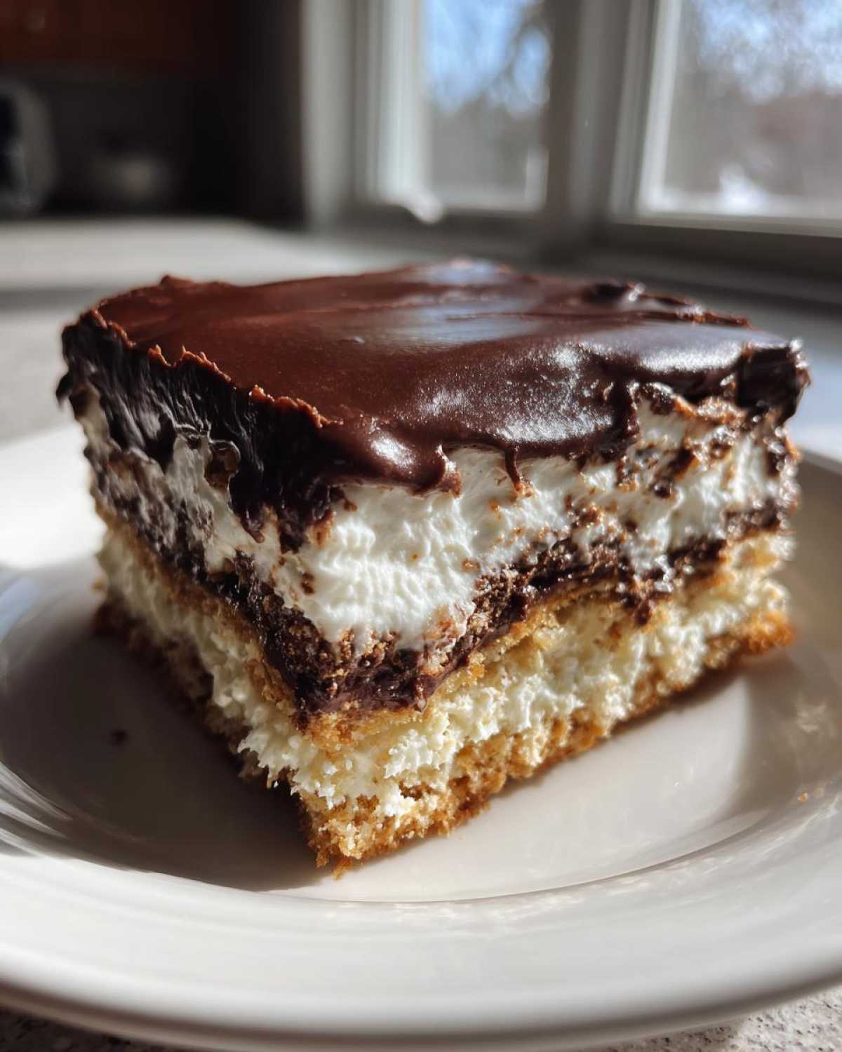 A square slice of layered eclair cake with a graham cracker crust, cream filling, and chocolate glaze.