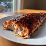 A close-up of a perfectly cooked, flaky salmon fillet with a dark, shiny glaze, resting on a white plate.