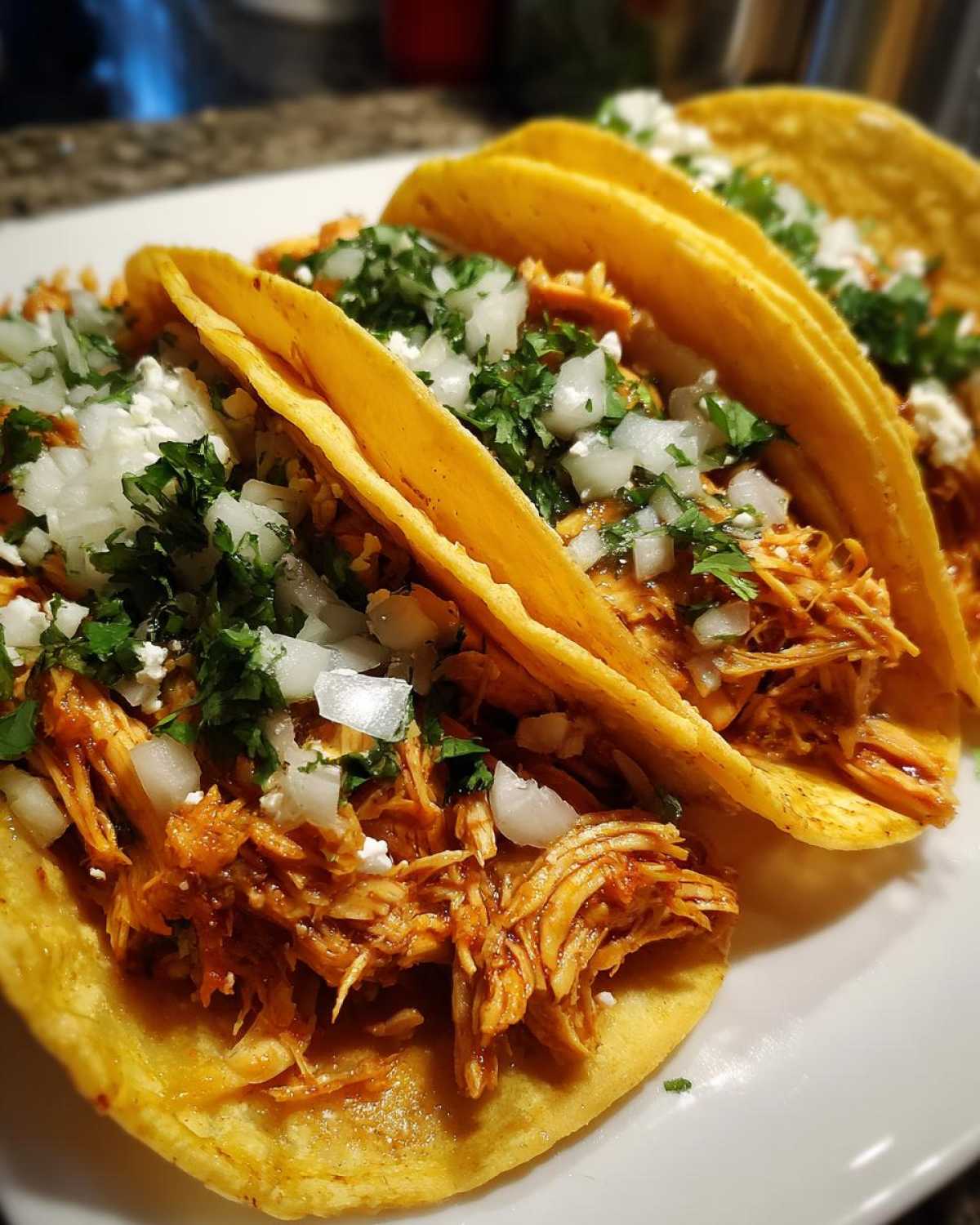 Close-up of three soft corn tortillas filled with shredded, saucy chicken, topped with diced white onion, cilantro, and cotija cheese for delicious chicken tacos.