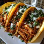 Three crispy shell chicken tacos filled with saucy shredded chicken, topped with diced white onion and fresh cilantro.