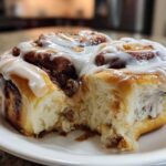 A close-up, featured image of a soft, fluffy cinnamon roll generously topped with white icing, with a piece already taken out.
