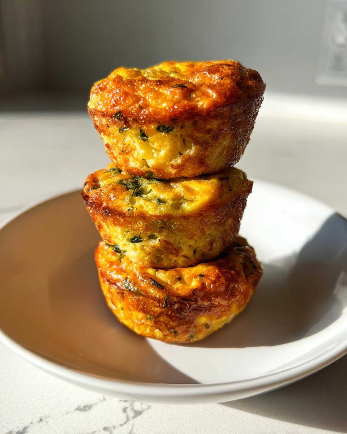 Three fluffy muffin tin egg bites stacked vertically on a white plate, showing golden-brown tops and visible green herbs.