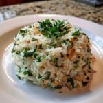 A mound of fluffy white rice pilaf mixed with herbs and toasted bits, served on a white plate.