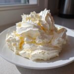 A generous mound of freshly whipped vanilla buttercream frosting sitting on a white plate, highlighted by natural light.