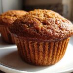 Close-up of two golden-brown, freshly baked honey bran muffins sitting on a white plate.