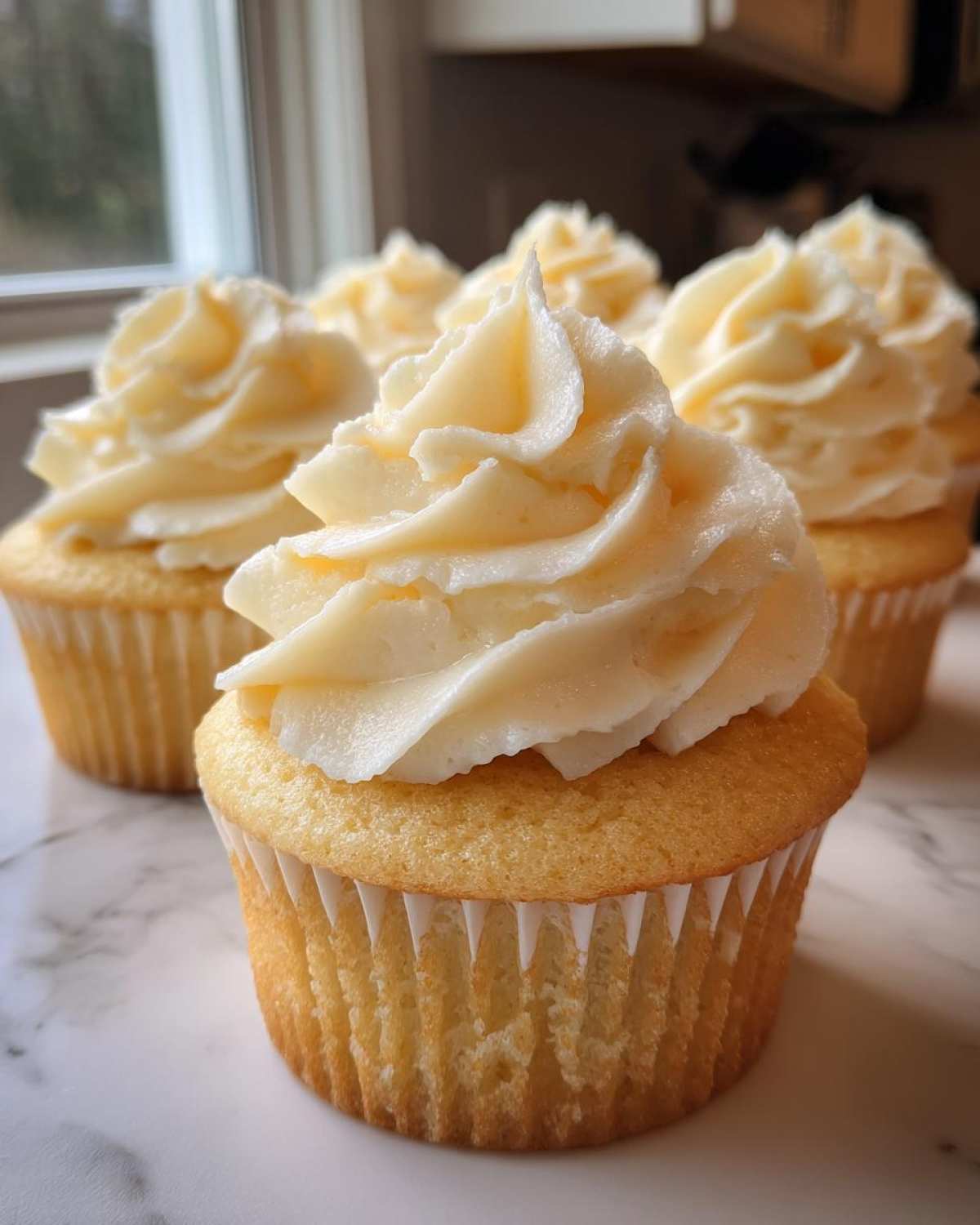 A close-up of several freshly baked vanilla cupcakes topped with thick swirls of creamy white frosting.