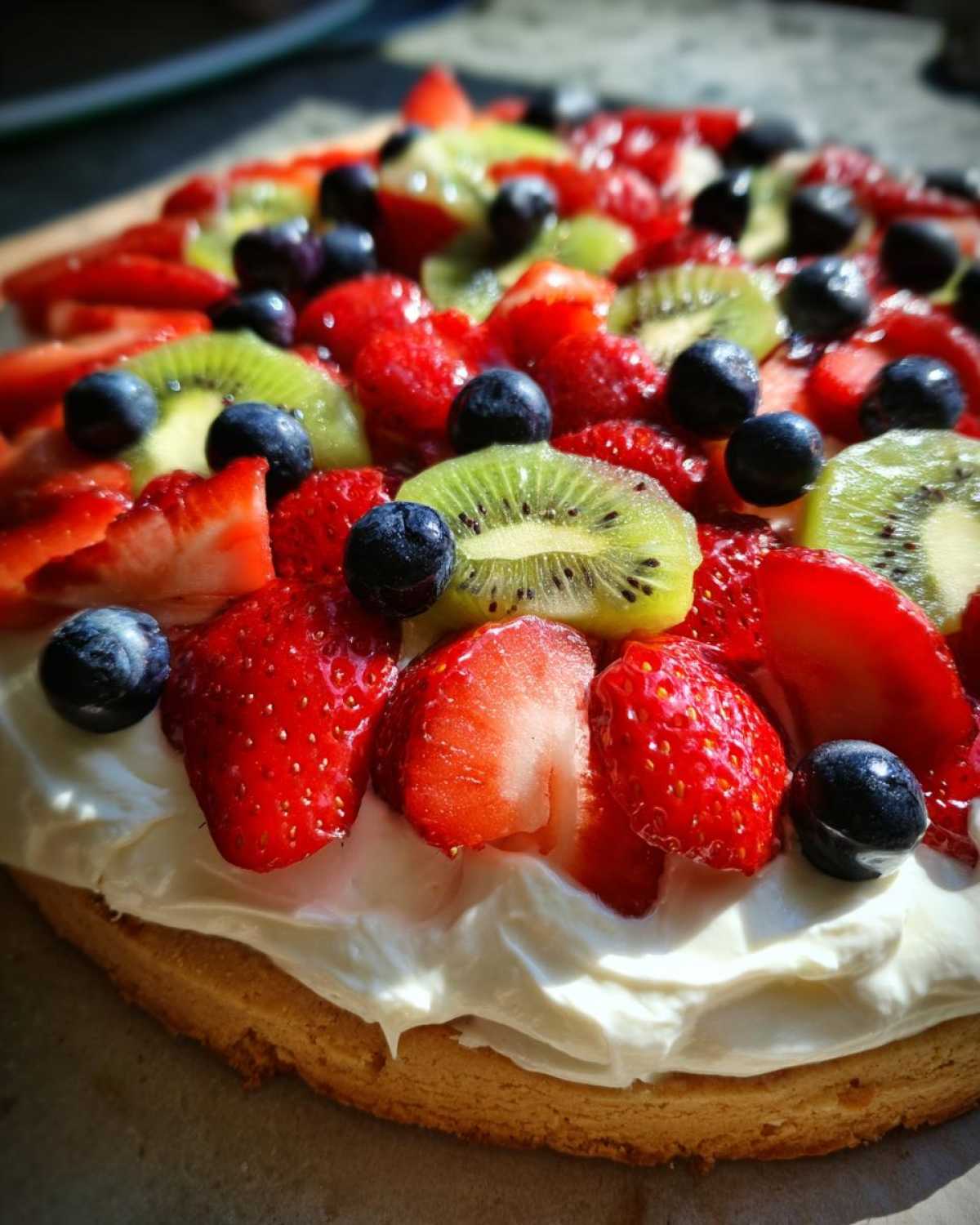 Close-up of a vibrant fruit pizza sugar cookie topped with cream cheese frosting, sliced strawberries, kiwi, and blueberries.