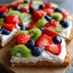 Close-up of a slice of fruit pizza sugar cookie topped with cream cheese frosting, strawberries, blueberries, and kiwi slices.