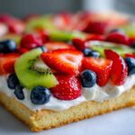 A close-up view of a slice of fruit pizza sugar cookie topped with cream cheese frosting and fresh berries.
