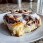 A close-up, appetizing shot of a freshly baked cinnamon roll slice generously topped with white icing.