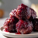 Close-up, appetizing shot of several Grape jelly meatballs piled on a small plate, dripping with thick, dark purple glaze.