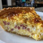 A close-up of a thick slice of Ham and Cheese Quiche with a golden-brown top, served on a white plate.