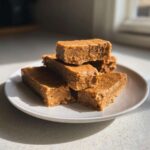 A stack of five homemade, dense, peanut-butter colored protein bars resting on a white plate.