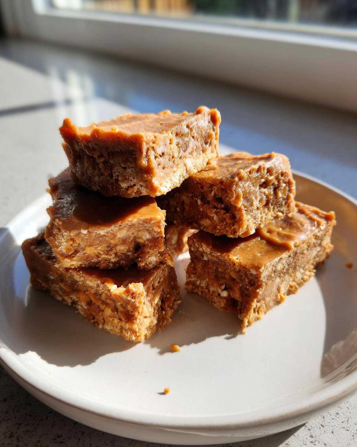 A stack of five homemade, peanut butter colored protein bars resting on a light ceramic plate near a window.
