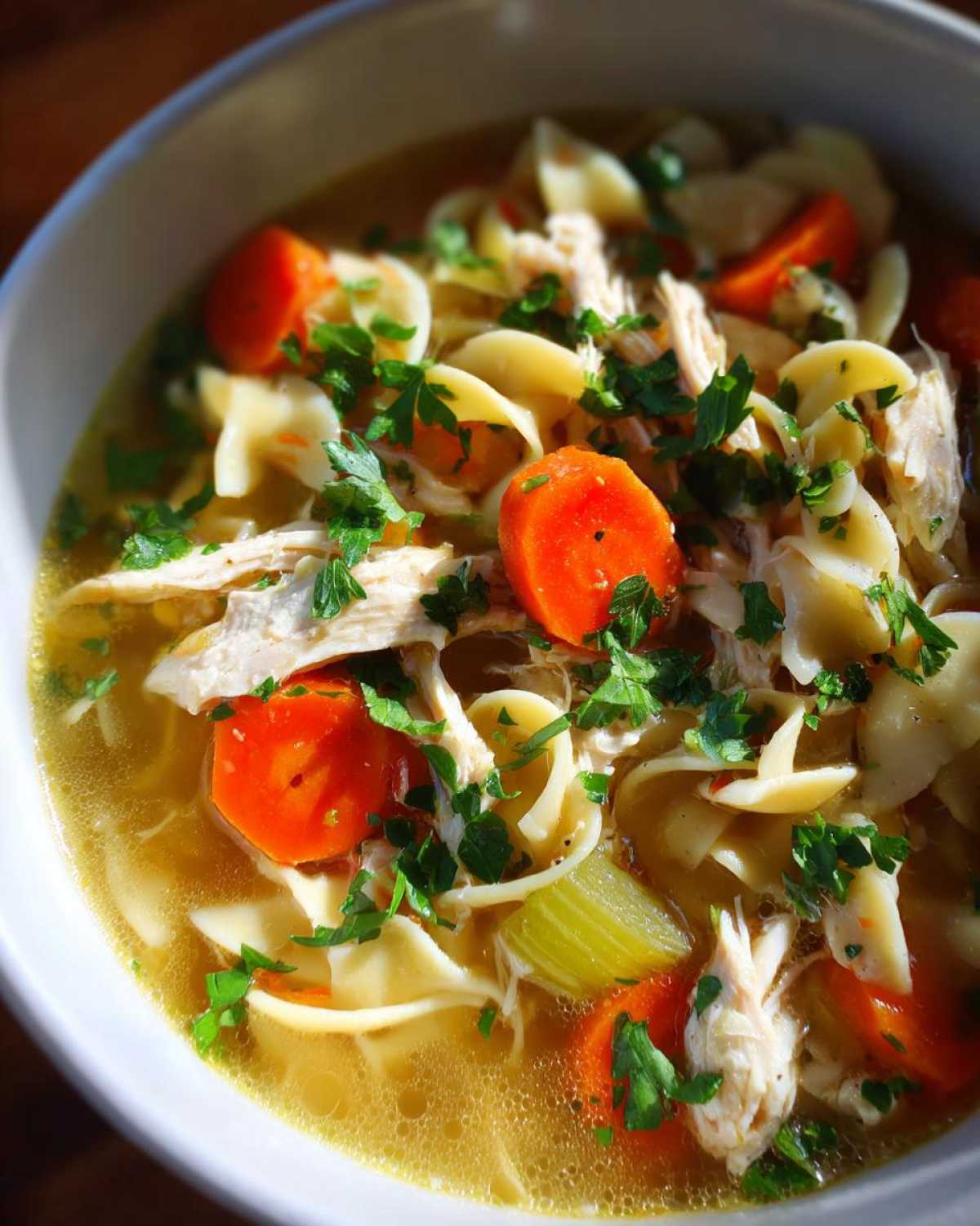 Close-up view of a hearty bowl of chicken noodle soup, featuring shredded chicken, bright orange carrots, egg noodles, and fresh parsley garnish.