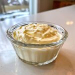 A small glass bowl filled with thick, creamy homemade mayonnaise resting on a white countertop.