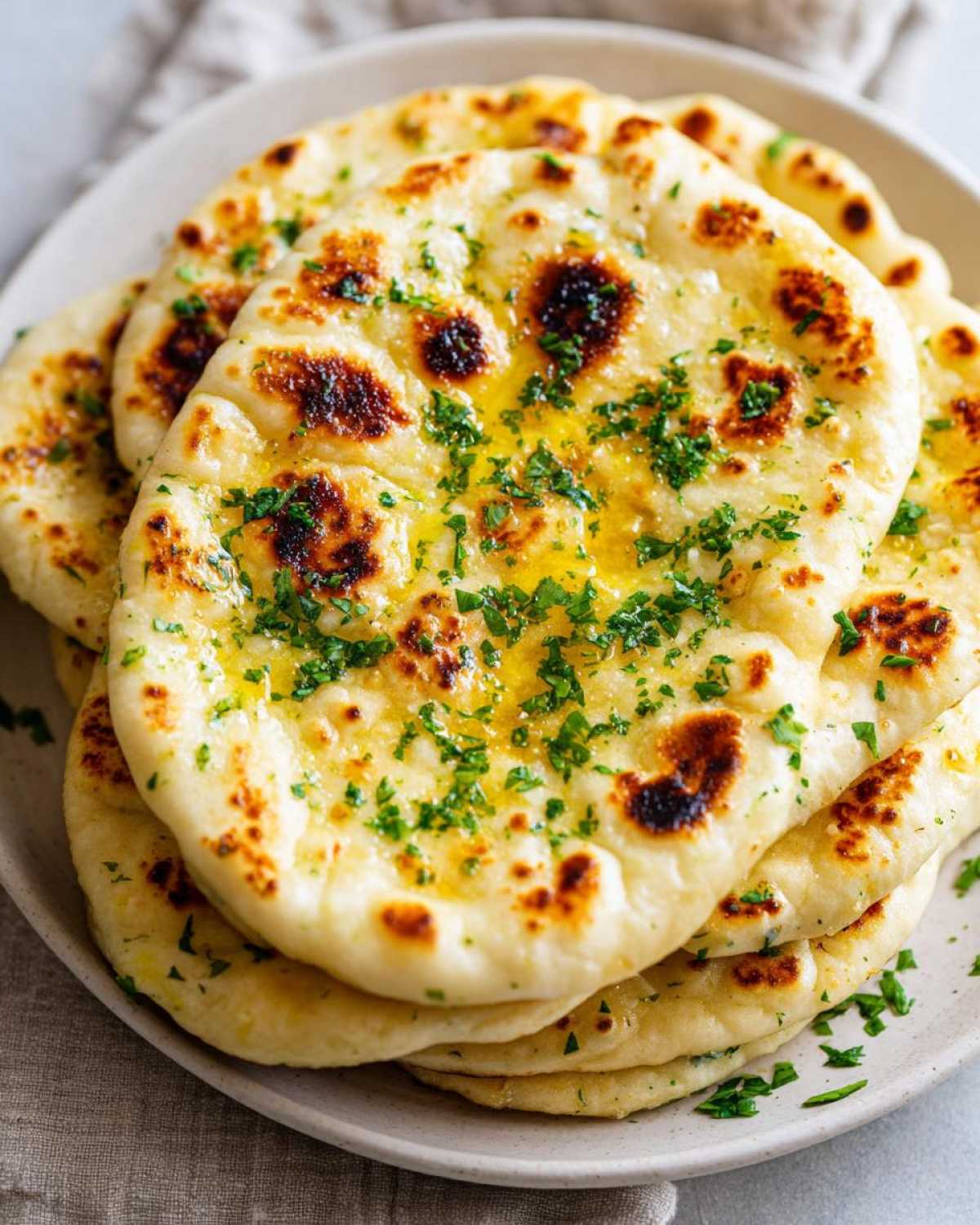 A stack of freshly made homemade naan bread brushed with melted butter and topped with chopped green herbs.