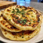 Close-up of a stack of three golden, fluffy homemade naan bread pieces topped with melted butter and fresh chopped parsley.