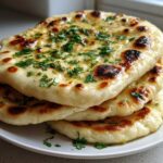 A stack of three fluffy, charred homemade naan bread pieces brushed with butter and topped with fresh green herbs.