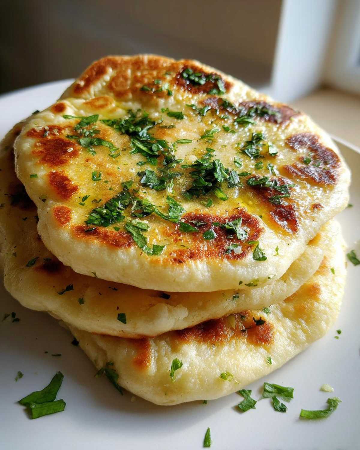 A stack of three fluffy homemade naan bread pieces, brushed with butter and topped with fresh chopped cilantro or parsley.