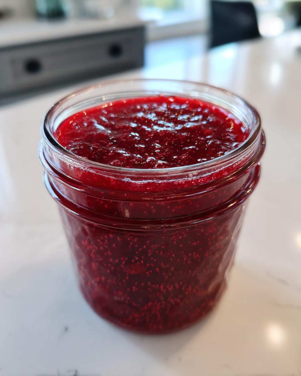 Close-up of a glass jar filled with vibrant, deep red homemade raspberry jam, showing visible seeds.