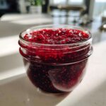 Close-up of a glass jar filled with vibrant, chunky homemade raspberry jam, catching the sunlight.
