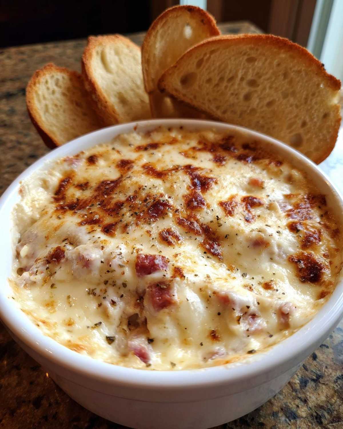 Close-up of a bubbling, cheesy Reuben dip in a white bowl served with toasted baguette slices for dipping.