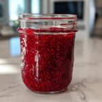 A full mason jar filled with vibrant, deep red homemade raspberry jam, showcasing many visible seeds.
