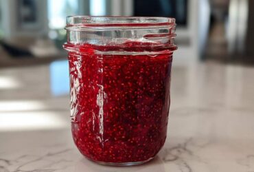 A full mason jar filled with vibrant, deep red homemade raspberry jam, showcasing many visible seeds.