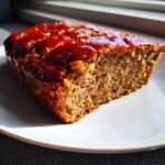A close-up of a juicy slice of Keto meatloaf topped with a shiny red glaze, sitting on a white plate.