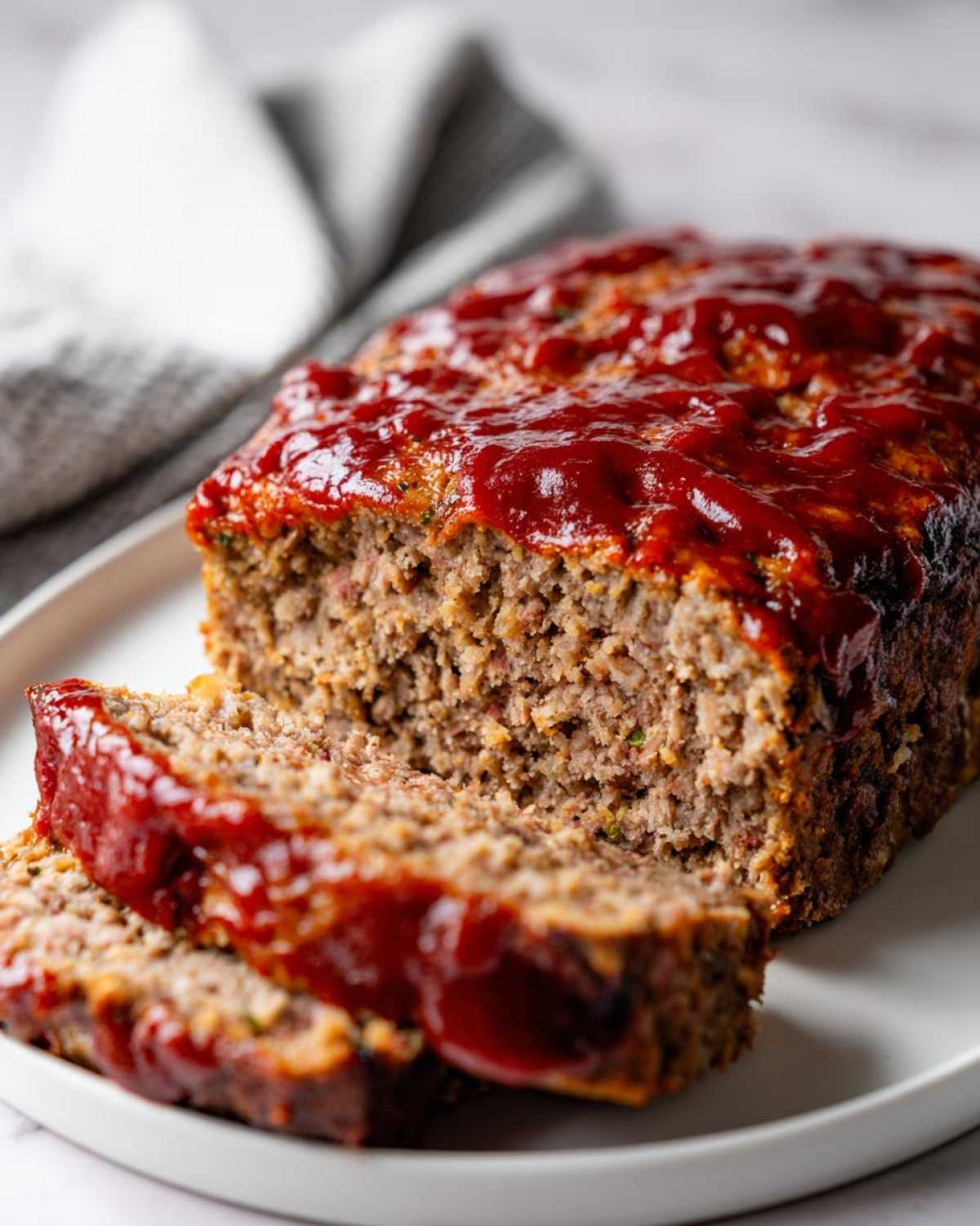 A freshly baked meatloaf, partially sliced, showing a moist interior and topped with a thick, glossy ketchup glaze. This is the perfect meatloaf.