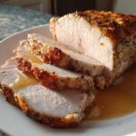 Close-up of a juicy pork loin roast, partially sliced, resting in its own pan juices on a white plate.