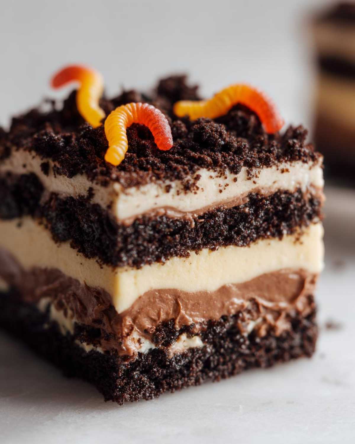 A square slice of layered dirt cake featuring chocolate cake, cream filling, and topped with crushed cookies and gummy worms.