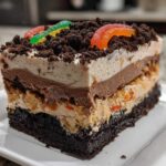 Cross-section slice of a layered dirt cake featuring a brownie base, pudding layers, and crushed cookie topping with gummy worms.